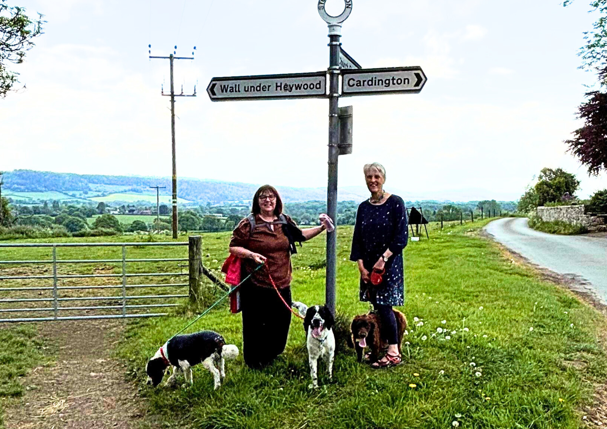 Two women and three dogs on leads ready for walk. Stood on grass verge one leaning on crossroad sign post and open countryside and hills in background. 