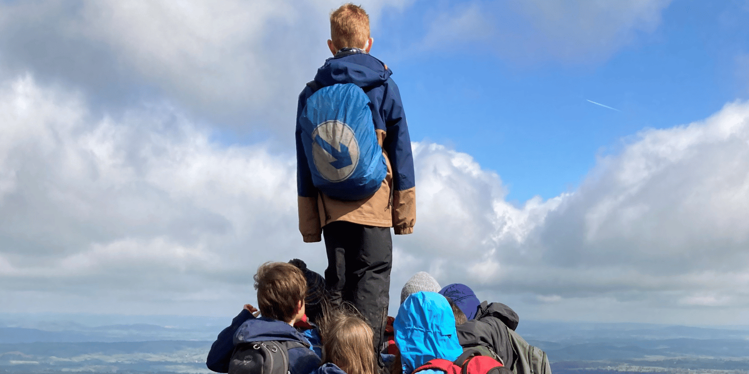 Boy stood ontop of cairn with his back to the camera wearing blue rucksack with white arrow 