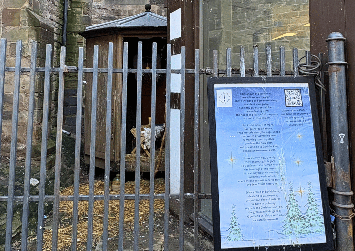 Railings with a wooden building behind, showing door open and a manger crib with straw